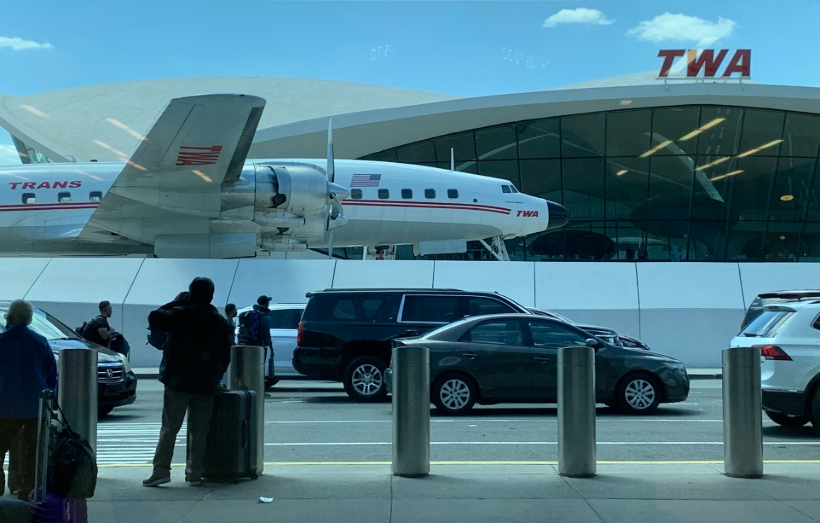 JFK car service vehicles at Terminal 5 pickup area, with historic TWA Hotel and aircraft in background.